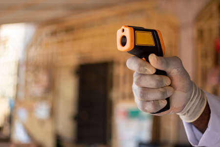 black person hand, wearing a glove and lab coat pointing an infrared thermometer, with blurred...