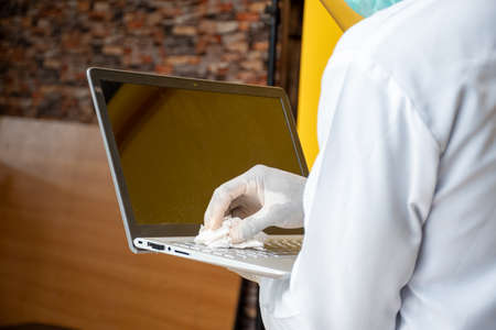 young black man wearing a lab coat and face mask and shield cleaning his laptop computer surface