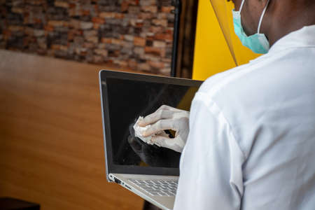 a young african man wearing a lab coat and face mask and shield cleaning his laptop computer surfaceの写真素材