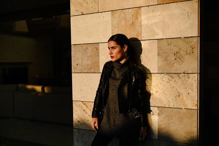 Young beautiful brunette girl posing at sunset on a wall background on a city street