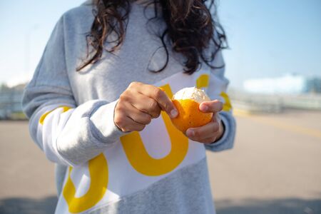 girl in sports hoodie hands removes the peel from an orange.の写真素材