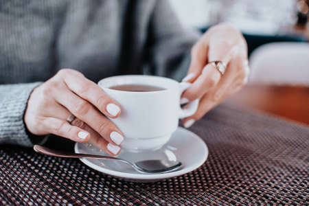 womens hands in a gray sweater with a white manicure hold a white mug of tea at a wooden table