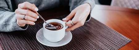 womens hands in a gray sweater with a white manicure hold a white mug of tea at a wooden tableの写真素材