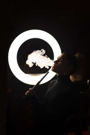 free woman smokes a hookah in the semi-darkness against the background of a ring lamp.