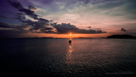 A serene sunset at Klong Muang Beach in Krabi, Thailand, featuring a boat on calm waters, silhouetted against a vibrant sky with clouds and distant islands.の写真素材