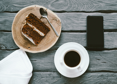 Piece of a homemade biscuit cake and black coffee on a black wooden table. Smartphone on a table with meal. Delicious breakfast of a busy person.の写真素材