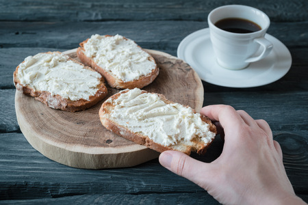 Hand holds homemade sandwiche with cream-cheese over a black wooden table. Wooden stand with sandwiches. Breakfast with black coffee.の写真素材