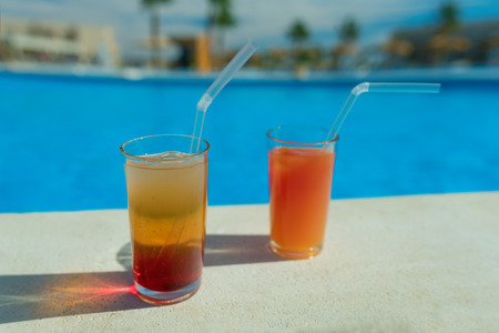Glasses with tasty cocktail in front of a swimming pool with clear water. Close-up photo. Palm trees on the background. Vacation at a resort in summerの写真素材