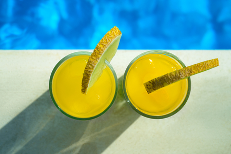 Glasses with yellow and green cocktail in front of a swimming pool with clear water. Close-up photo. Top view at glasses with cocktail and watermelon.の写真素材