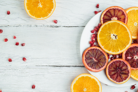 Slices of sicilian orange and common orange on a white wooden table. Pomegranate seeds scattered on a table. Top view. Space for text.の写真素材