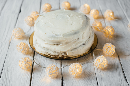 Celebration homemade biscuit cake with white cream on a white wooden table. Twinkling lights around the cake.の写真素材