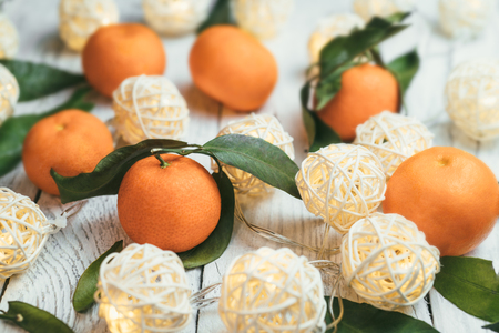 Mandarins with green leaves on a white wooden table. Twinkling lights on a table. Tasty tangerines for the New Year festive tableの写真素材