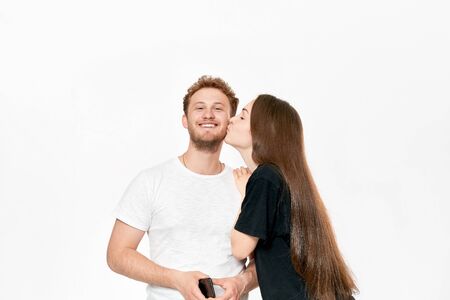 Studio shot of a happy couple kissing. Girlfriend kisses boyfriend's cheek. He smiles pleased while standing infront of white background.の写真素材