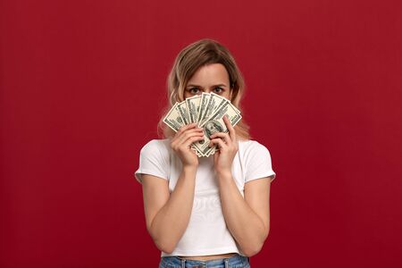 Portrait of a girl with curly blond hair dressed in a white t-shirt standing on a red background. Happy model holding American dollars.の写真素材