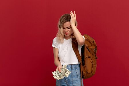 Portrait of a girl with curly blond hair, orange backpack and white headset dressed in a white t-shirt standing on a red background expressing puzzled emotion because she doesn't have enough money.の写真素材