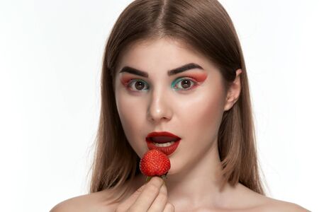 Closeup portrait of a surprised beautiful young woman with bright color make-up holding strawberry near the face. Isolated over white background.の写真素材