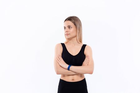 Young sporty blond woman in a black sportswear with smart watches for pulse measuring keeping hands crossed standing over white background looking to the left. Place for advertisement.の写真素材