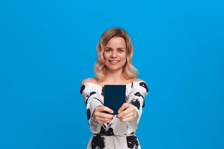 Pretty blond young woman in a white dress with flowers holding passport and smiling to the camera....