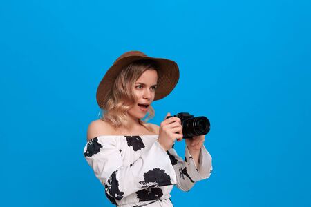 Portrait of a girl with curly blond hair in a sundown hat and white dress stands on a blue background. Model takes photos with retro camera.の写真素材
