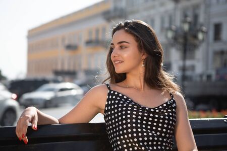Beautiful young woman in a black dress sits on a bench resting after sightseeing tour in old European town.の写真素材