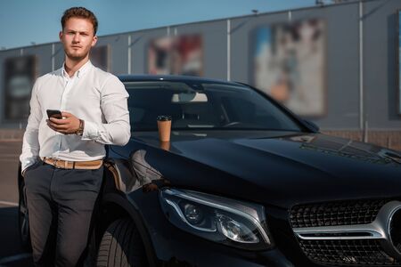 Young successful businessman in a white shirt checking smartphone standing near his luxary car. Disposable cup of hot coffee on a motor hood. Driver takes a break to drink coffee.の写真素材