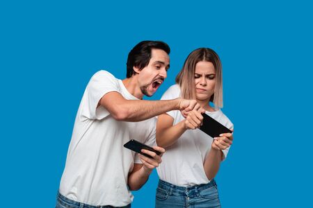 Beautiful couple wearing white t-shirts standing isolated over blue background, playing games on...