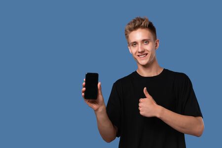 Blond teenager in a black tee holding smartphone with screen to the camera with thumb up isolated...