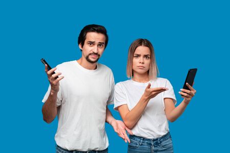Brunet man in white tees and blue jeans is shocked looking at the smartphone screen of a blond...