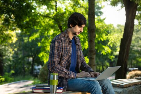 Young brunet freelancer with beard and mustache sitting with a laptop and coffee in a park. Work at any placeの写真素材