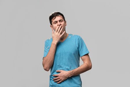 Young brunet man with white feather from a pillow yawning isolated over grey background. Being tired...