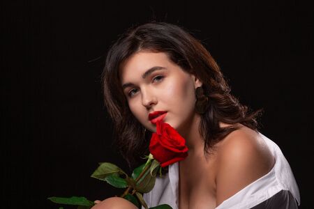 Beautiful woman with bright makeup posing isolated over black background with red rose. Image of young beautiful woman with red lips in a white shirtの写真素材