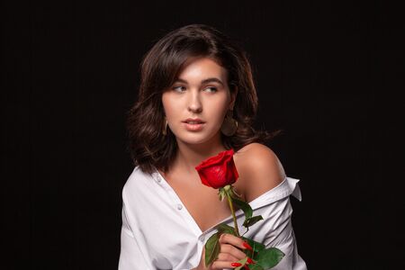Beautiful woman with bright makeup posing isolated over black background with red rose. Image of young beautiful woman with red lips in a white shirtの写真素材