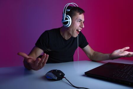 Angry gamer wearing headphones playing video games on computer in a dark room lit with neon lights. Video games championshipの写真素材