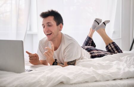 Handsome young man in the white shirt and pajama on a bed with white linen is excited looking at the...