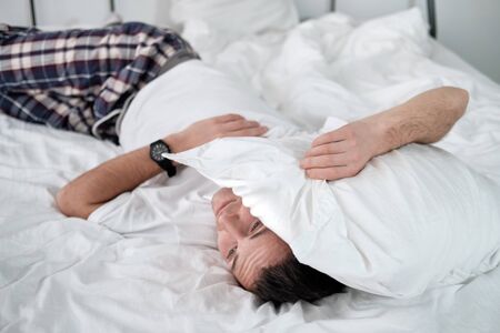 Young man in the white shirt lying on a bed with white linen and covers his head with a pollow....