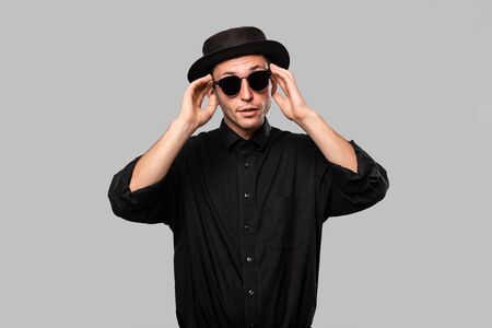 Portrait of a stylish handsome man in a black shirt, pork pie and sun glasses hat isolated over grey background.の写真素材