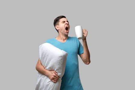 Young brunet man yawning holding white cup of coffee and white pillow. Waking up in the morning.の写真素材