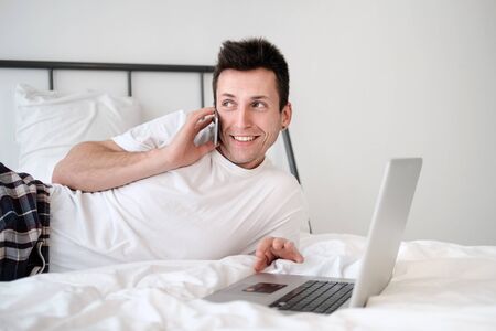 Young man in the white shirt and pajama sitting on a bed looking at the screen holding bank card. Online shoppingの写真素材