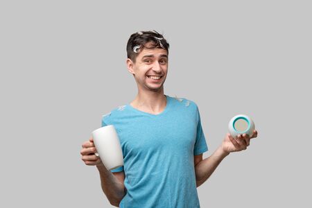 Young brunet man holding white cup and alarm-clock isolated over grey background. White feather from a pillow on the mans head. Time to wake upの写真素材