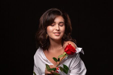 Beautiful woman with bright makeup posing isolated over black background with red rose. Image of young beautiful woman with red lips in a white shirtの写真素材