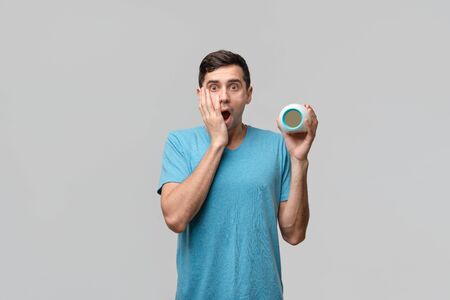 Young brunet man is shocked holding alarm-clock isolated over grey background. Time to wake upの写真素材