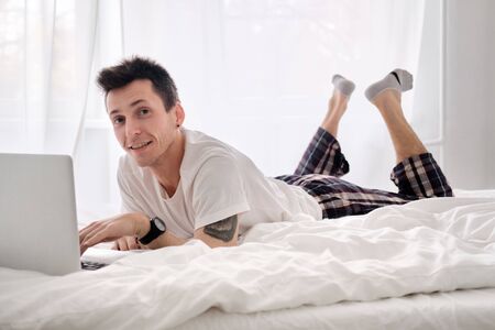 Handsome young man in the white shirt and pajama on a bed with white linen is excited looking at the screen of a laptop. Morning of a freelancerの写真素材