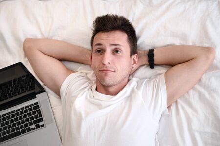 Young man in the white shirt smiling lying on a bed with white linen. Concept of relaxation. Laptop is next to a manの写真素材