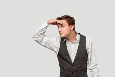 Handsome young man in a white shirt and grey vest holds palm of a hand over his eyes to see better.の写真素材