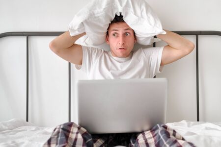 Angry handsome young man in the white shirt and pajama holding white pillow on a head and laptop on kneesの写真素材
