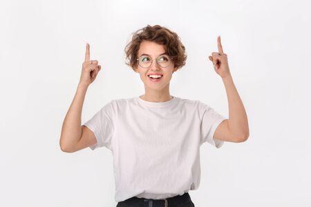 Pretty smiling woman in white shirt and eyeglasses pointing up at copy space. Place for advertisingの写真素材