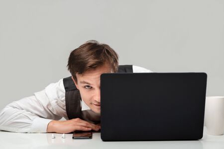Businessman in a white t-shirt and grey vest is hiding behind laptop. Having problems. Daily business routine.の写真素材