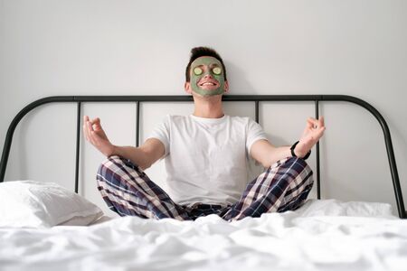 Young man in a white shirt with applied green cosmetic mask and pieces of cucumber on eyes is relaxing on a bed with white bed linen. Concept of wellbeing and facial treatment for menの写真素材