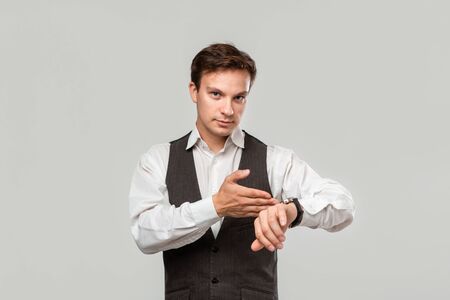 Young businessman in a white shirt and grey vest pointing at the hand watch to highlight that tie has come. Concept of timeの写真素材
