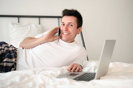 Young man in the white shirt and pajama sitting on a bed looking at the screen holding bank card. Online shoppingの写真素材